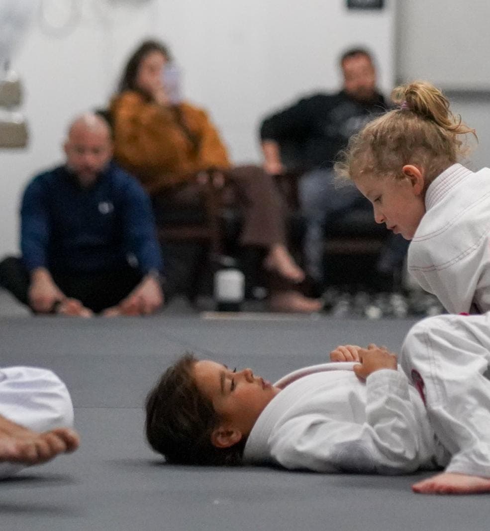 Two young students in white gis practice a technique on the mat while parents watch.